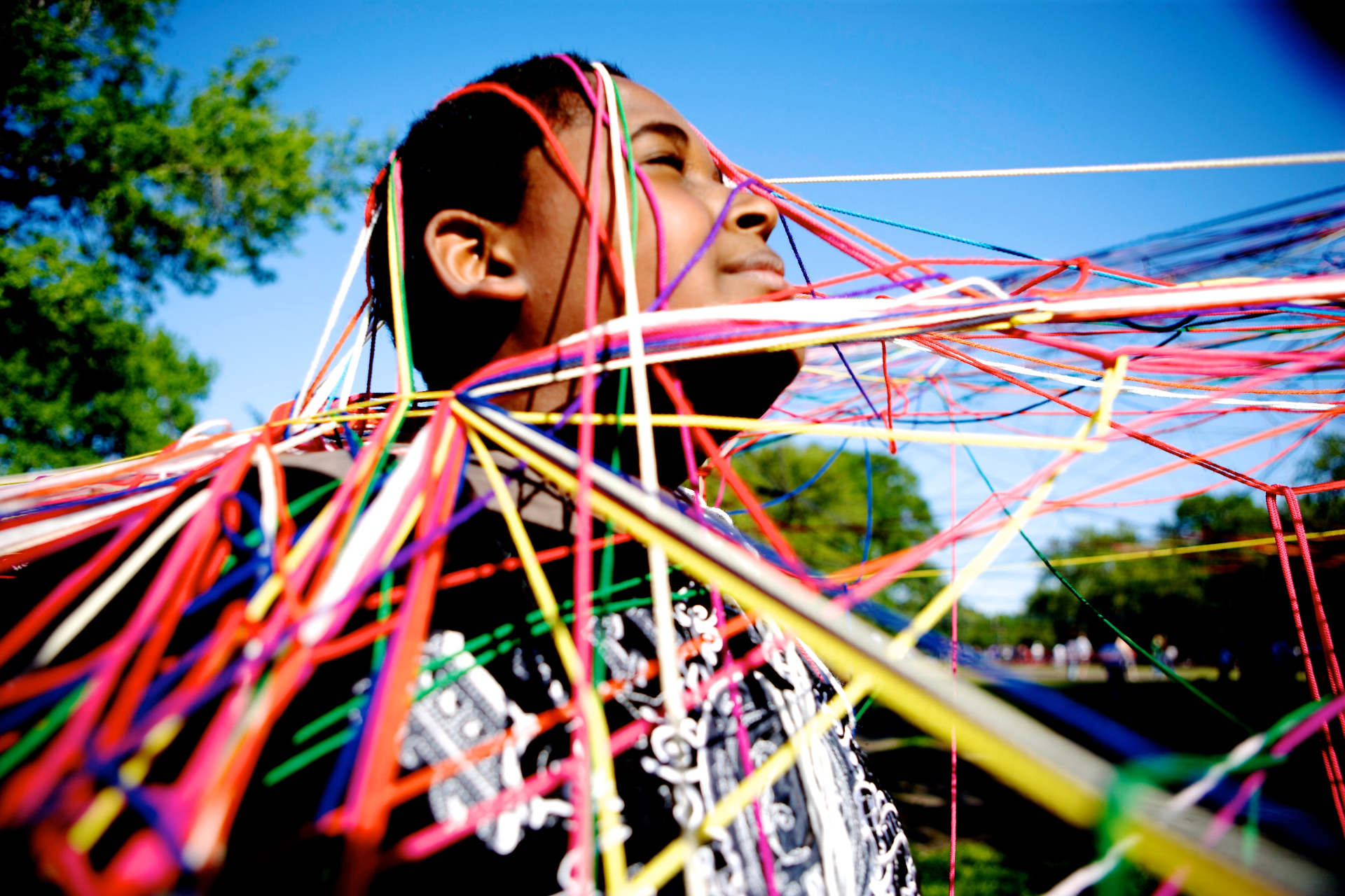 A child stands in the sun with their eyes closed, wrapped in a web of rainbow-colored strings from Jasmine Zimmerman's Collective String Web installation